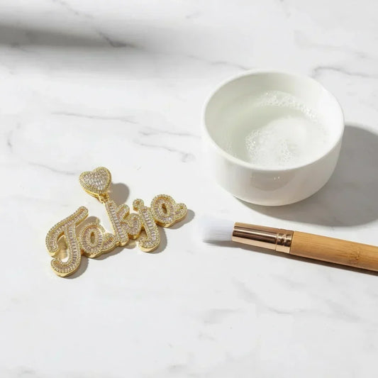 Gold iced-out baguette cursive name pendant beside a bowl of soapy water and soft brush — how to clean CZ jewelry safely at home