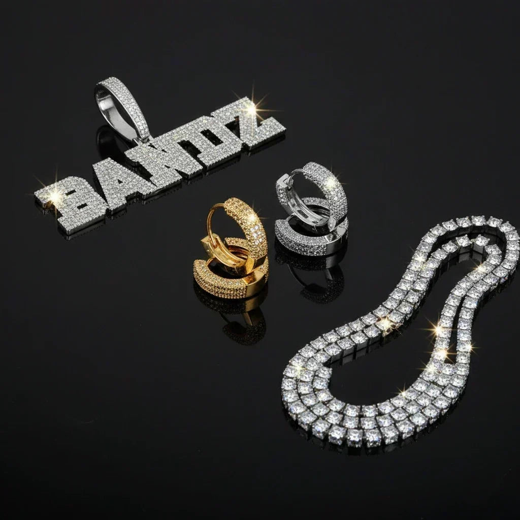 Iced out name pendant, gold and silver huggie earrings, and tennis chain flat lay on black background