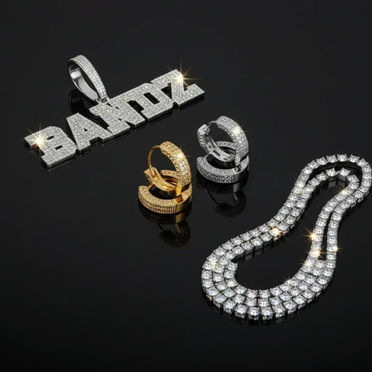 Iced out name pendant, gold and silver huggie earrings, and tennis chain flat lay on black background